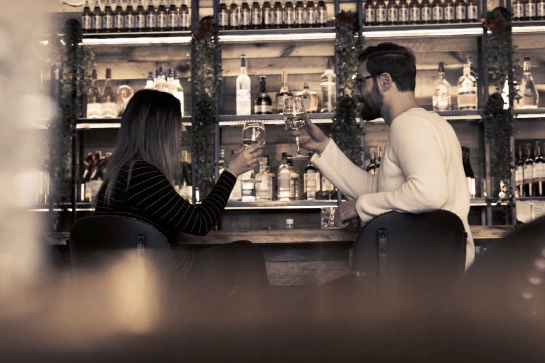 a man and a woman sitting at a bar cheersing over a glass of wine